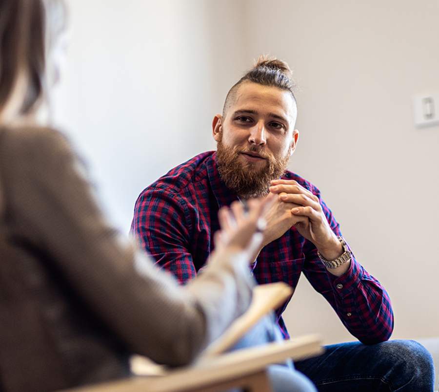 A man with a beard and a top knot, wearing a red and blue plaid shirt, attentively listens during an individual counselling session.