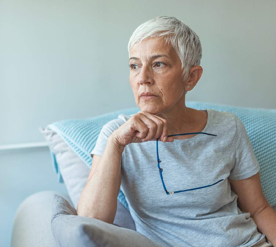 A mature woman with short grey hair sits on a couch, holding her glasses in one hand waiting patiently.