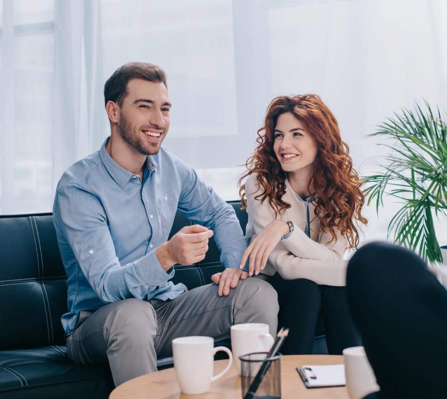 A smiling couple sitting on a sofa during a counseling session, relaxed and enjoying a warm conversation with a therapist over coffee.