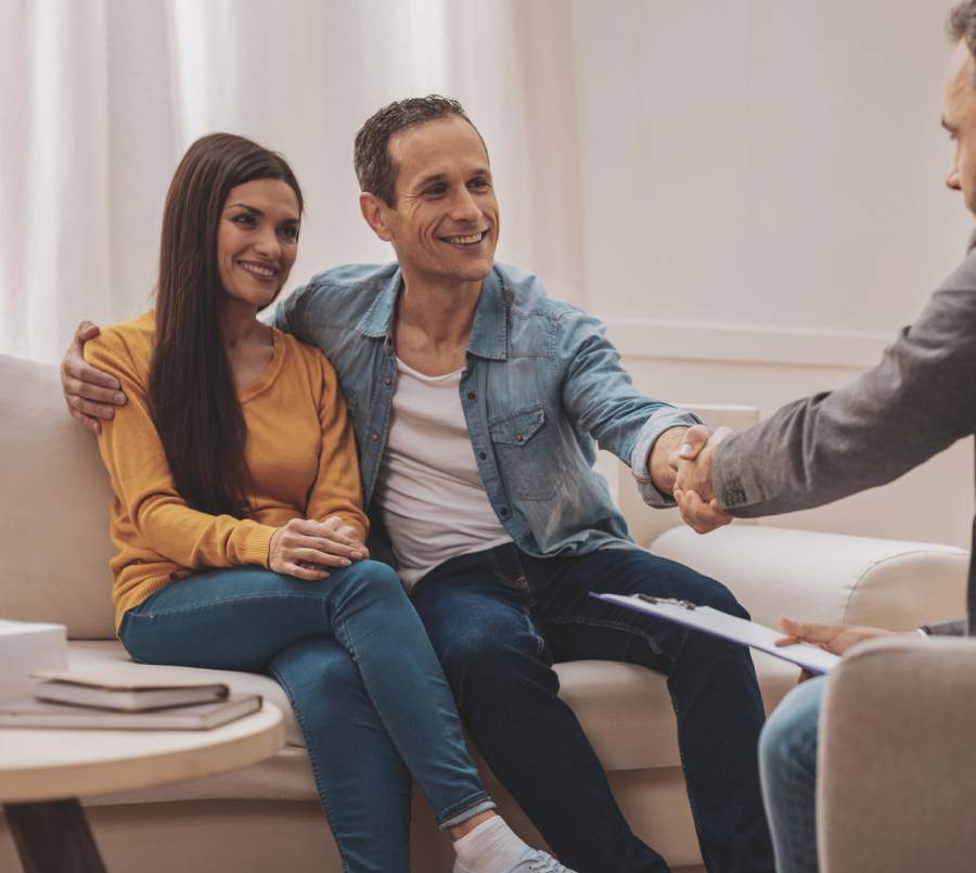 A happy couple sitting closely together on a couch, shaking hands with a therapist during a relationship counseling session.